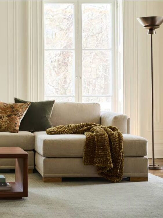 A beige couch with green and floral pillows, draped with a brown blanket. It's placed in front of a large window, next to a floor lamp, on a carpeted floor.