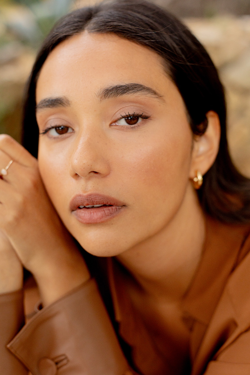 A person with long dark hair is looking at the camera. They are wearing a brown top and small hoop earrings.