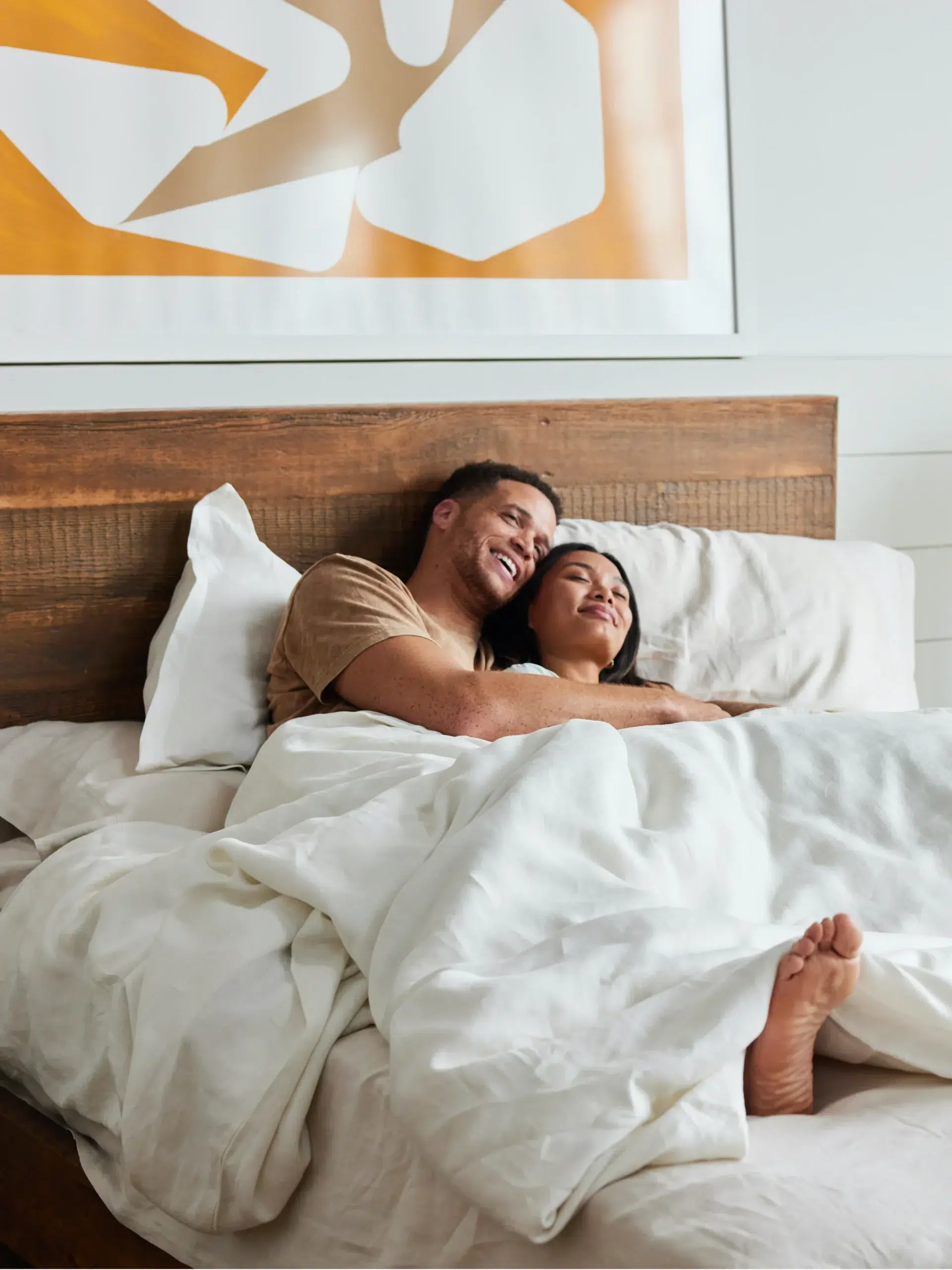 Two people lie in bed under white sheets. One is smiling and holding the other, who is resting with eyes closed. They are on a wooden bed with white pillows and a framed abstract artwork above.