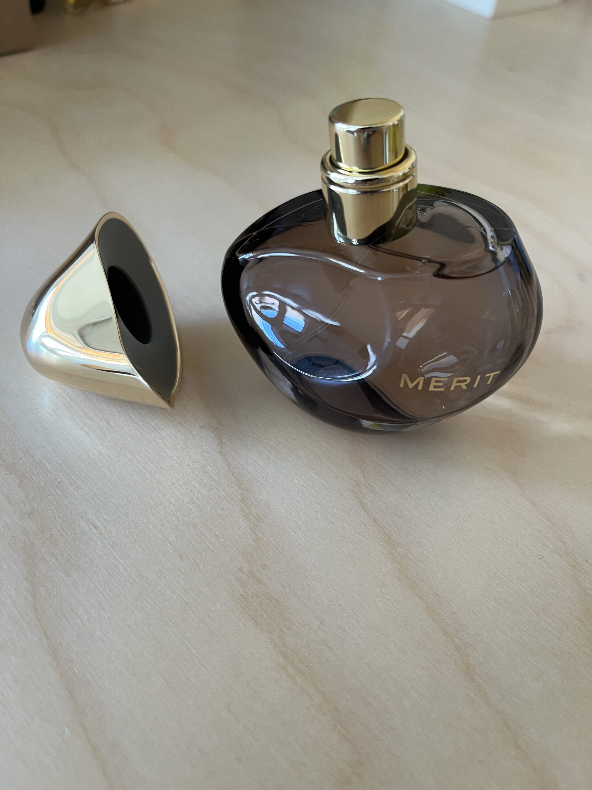 A dark glass perfume bottle labeled "Merit" with a gold cap removed, placed on a light-colored surface.