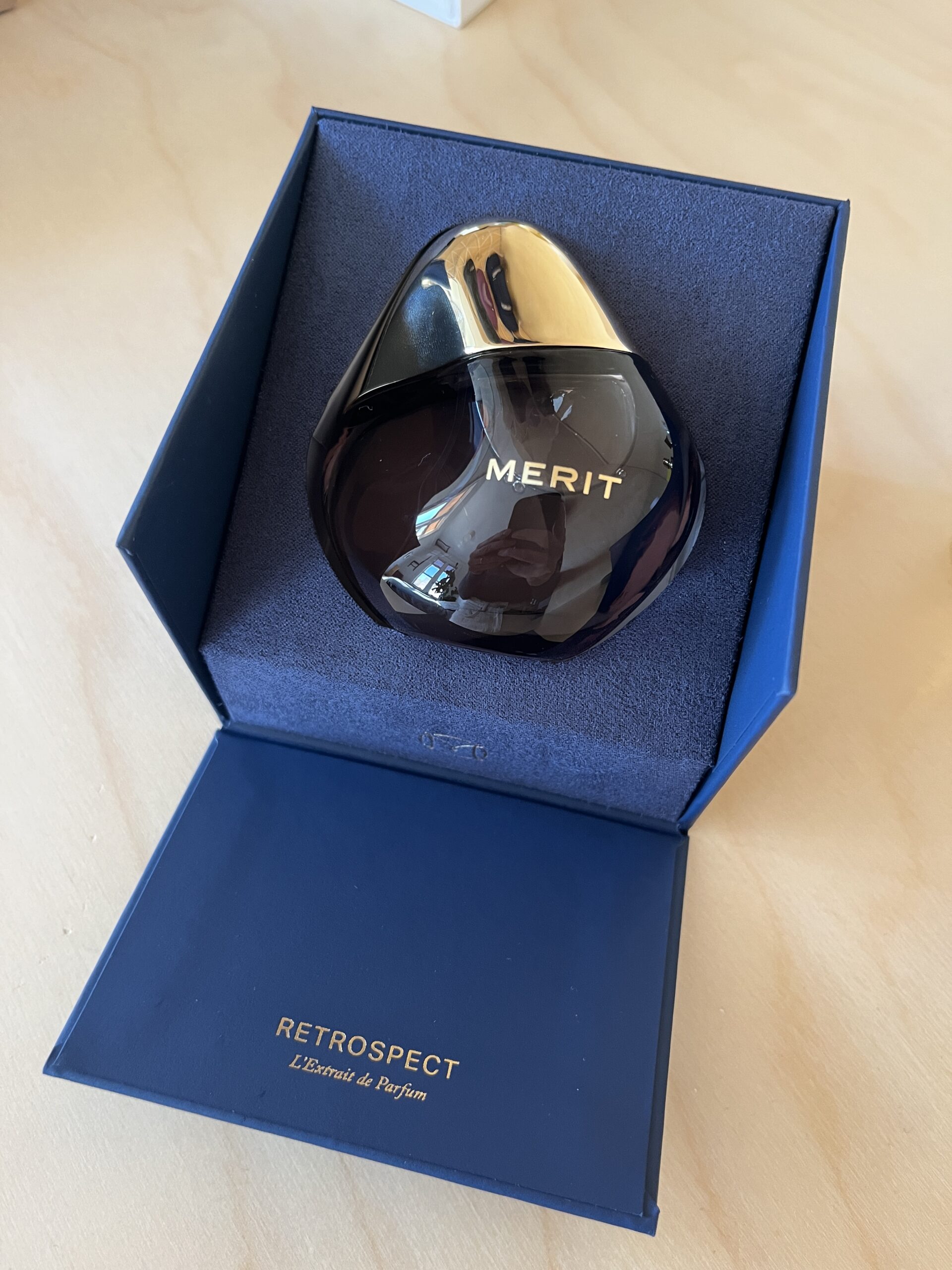 A teardrop-shaped perfume bottle labeled "Merit" inside an open blue box with "Retrospect L'Etoile de Parfum" printed on the lid.