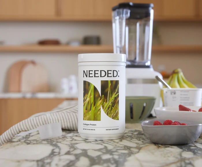 A white container labeled "Needed: Collagen Protein" on a kitchen countertop, surrounded by a blender and bowls of bananas and raspberries.