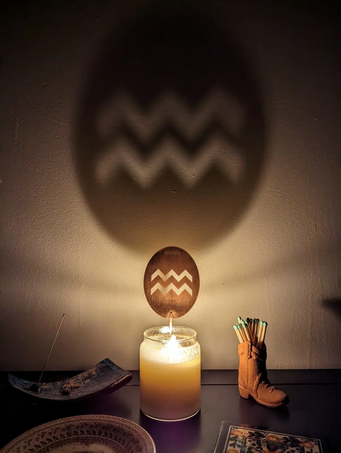 Candle with zigzag pattern casts shadow on wall; nearby, an incense holder, decorative boot with items, and a bowl.