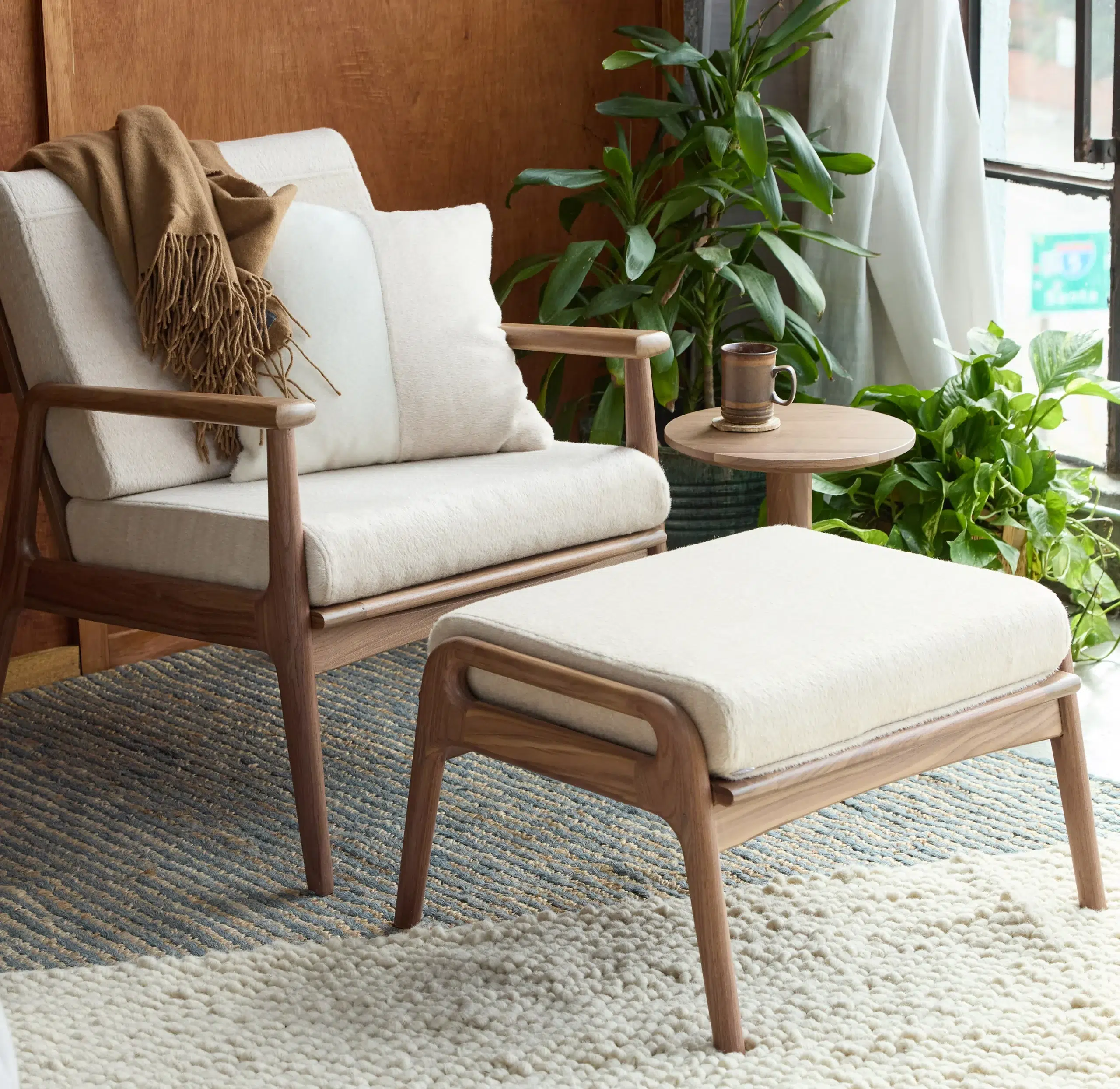 A cozy chair with a brown throw is next to a round side table holding a mug. A matching ottoman and various plants surround the setting on a textured rug by a window.