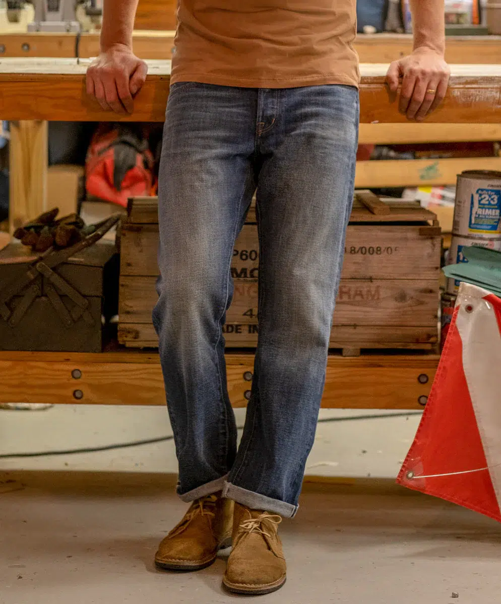 Person in blue jeans and brown shoes leaning against a wooden workbench in a workshop setting.