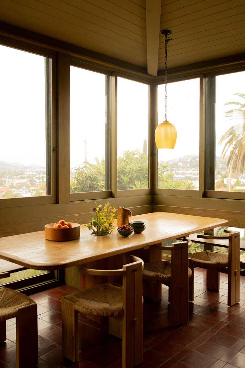 A wooden dining table with chairs is set by large windows. A bowl of oranges, a vase of flowers, and a pendant light add to the decor. Scenic views of trees and hills are visible outside.