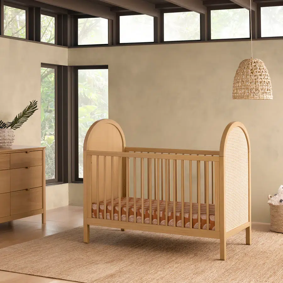 A wooden crib with a striped mattress is placed in a modern room with large windows, a dresser, and a woven light fixture.