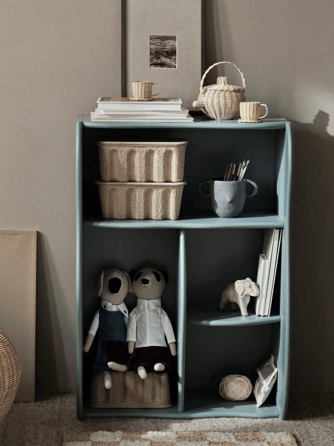 A small, light blue shelf with baskets, books, and toys sits against a beige wall. Two plush toys and an elephant figurine are on the lower shelf. A woven basket is on the left.