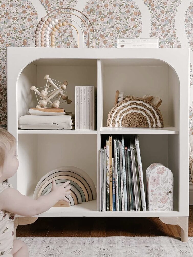 A toddler reaches for a wooden rainbow toy on a white shelf filled with books and toys, set against a floral wallpaper background.