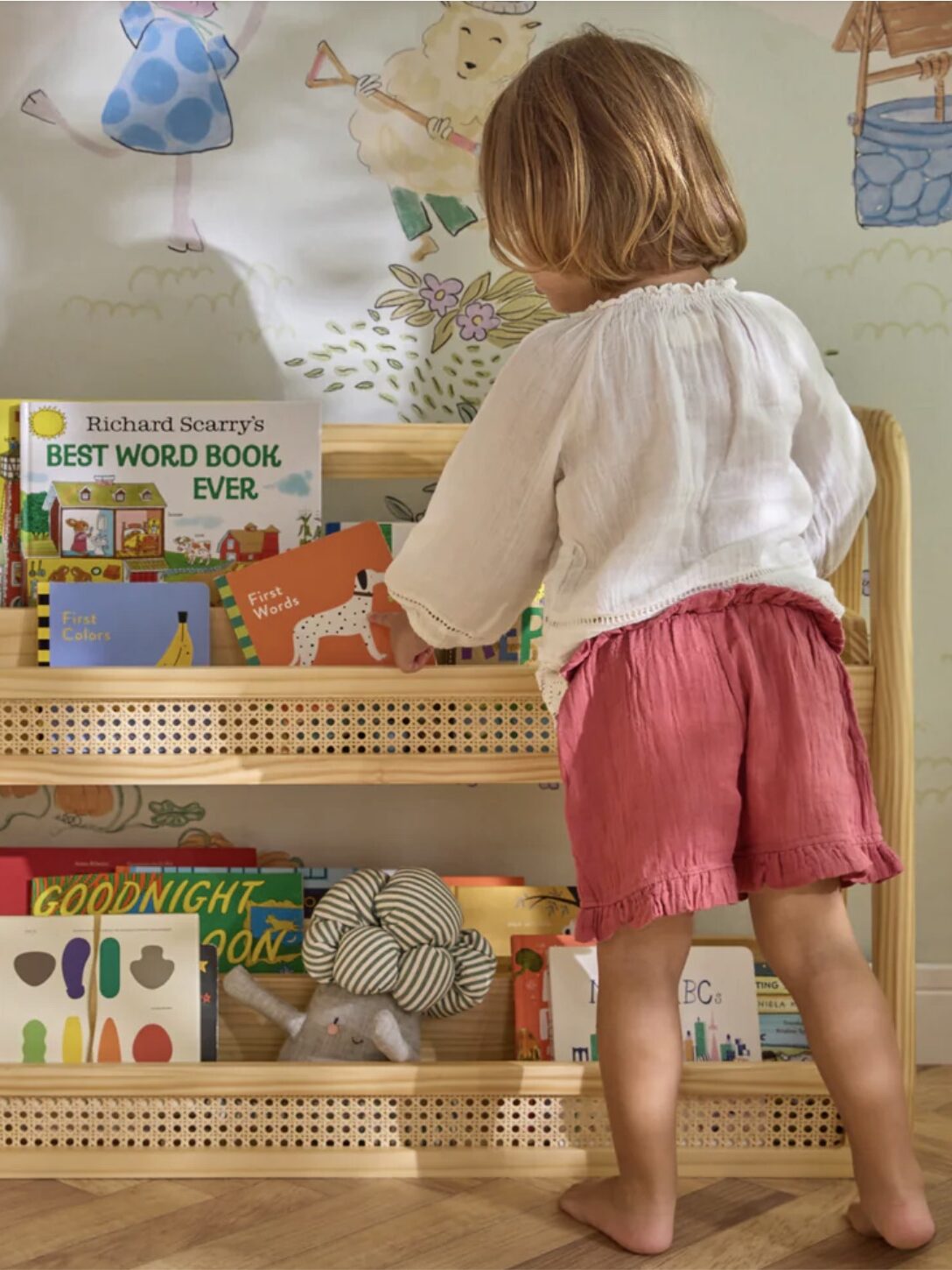 A child in a white blouse and pink shorts stands in front of a bookshelf filled with children's books and toys. A colorful mural decorates the wall behind.