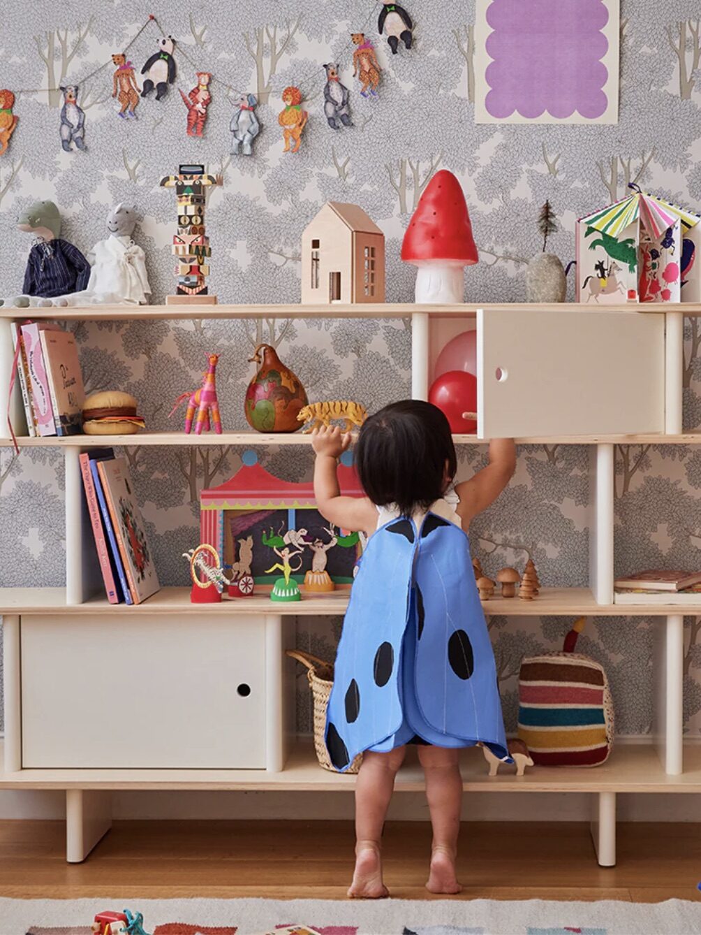 A child in a blue ladybug costume reaches for toys on a wooden shelf with various decorations in a playroom.