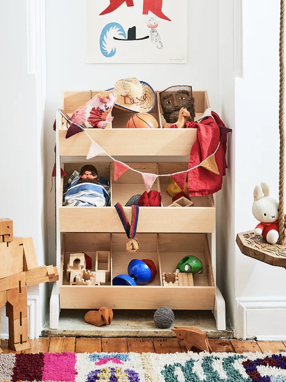 Wooden storage bins filled with toys, including a doll, basketball, stuffed animals, and a cowboy hat. A wooden robot stands nearby, and a rope swing hangs on the right.