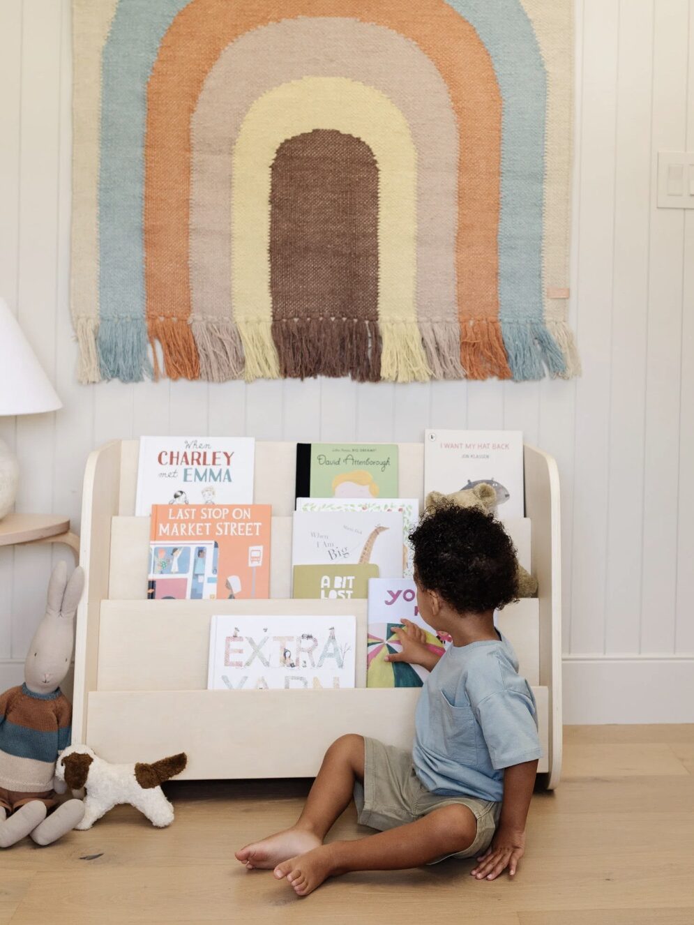 A child sits on the floor, reaching for a book from a wooden bookshelf filled with children's books. A colorful woven wall hanging is displayed above, and a stuffed rabbit toy is nearby.