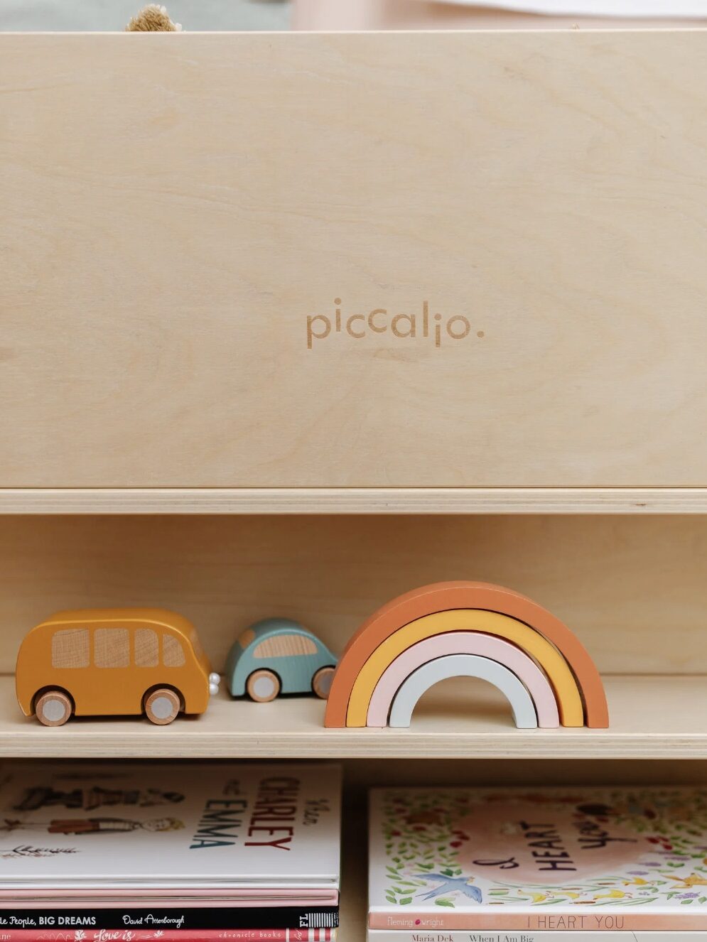 Wooden toy cars and a rainbow are displayed on a wooden shelf labeled "piccalio," with children's books underneath.