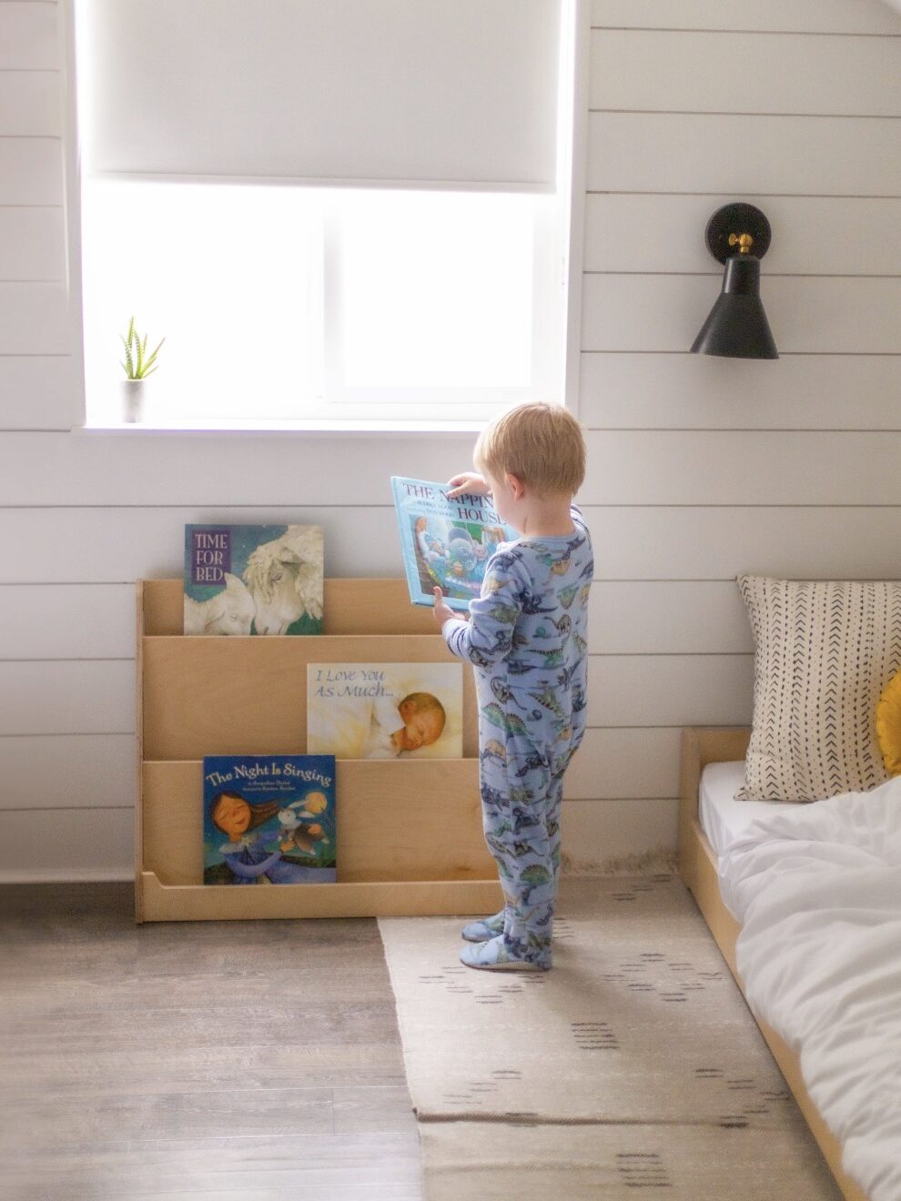 A child in pajamas stands in a bedroom holding a book in front of a window. Books are displayed in a wooden wall shelf. A bed is nearby, and a plant sits on the window sill.