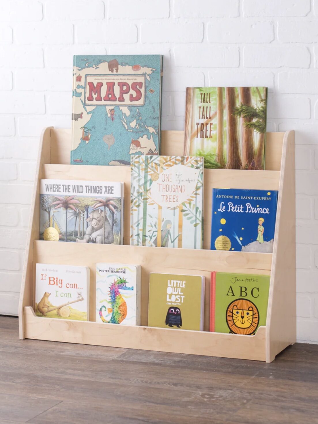 A wooden bookshelf displays children's books, including titles like "Maps," "Le Petit Prince," and "ABC." The shelf is against a white brick wall on a wooden floor.