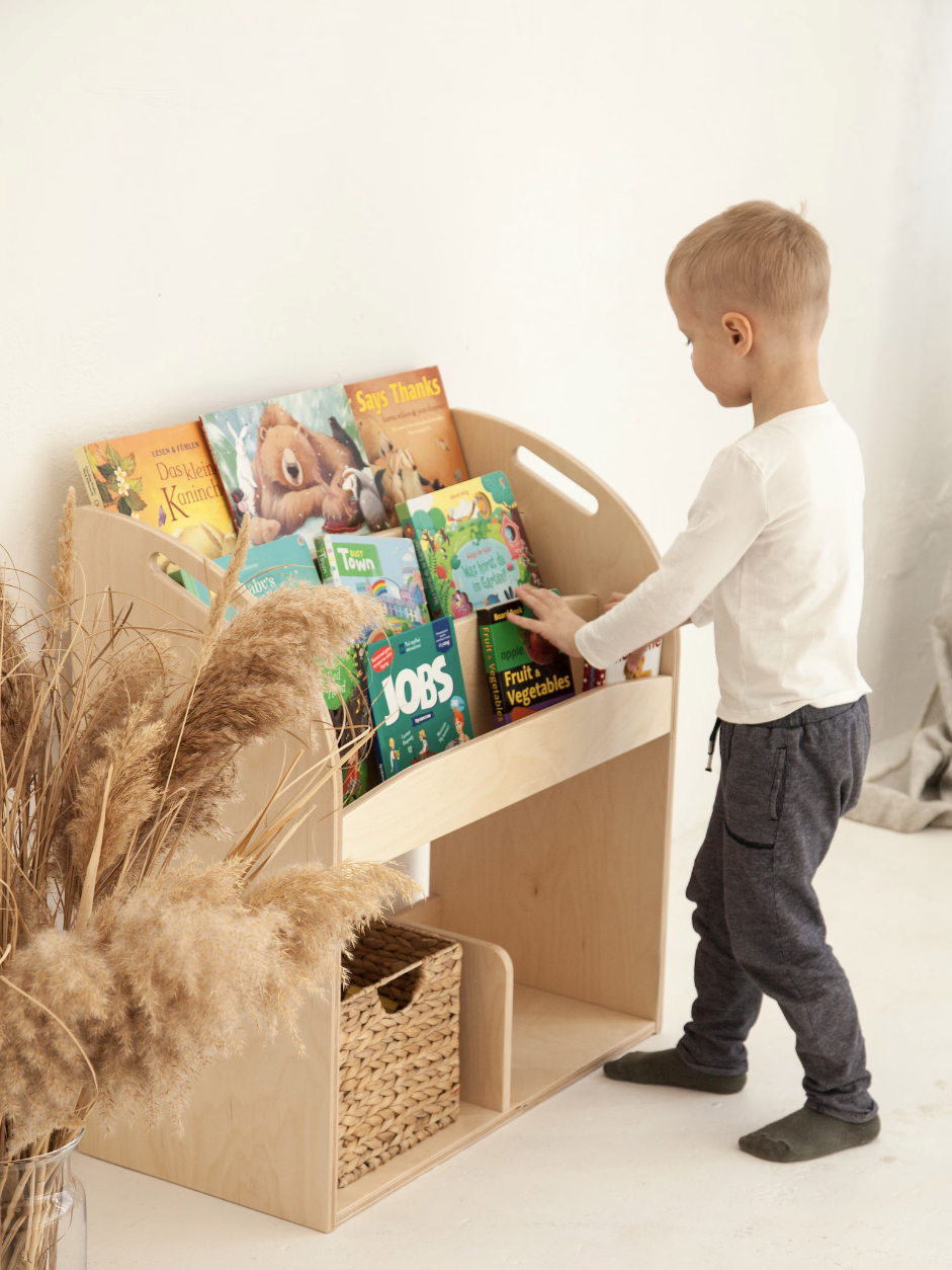 A child stands next to a wooden bookshelf filled with colorful books. Pampas grass in a vase decorates the floor.