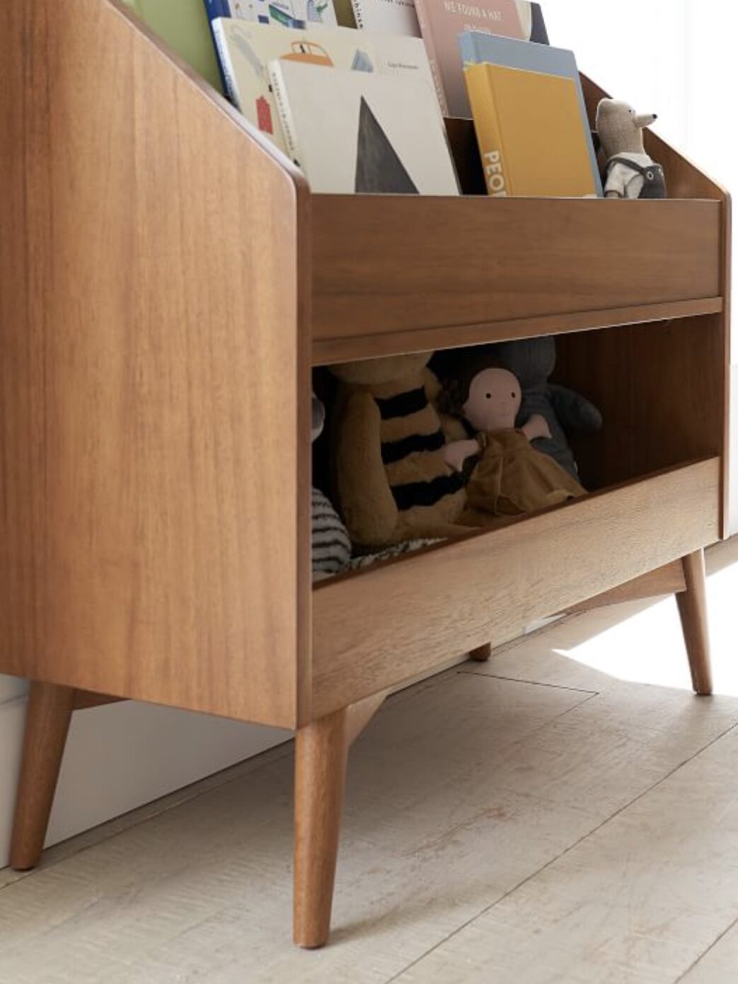 A wooden bookshelf with an upper shelf of books and a lower shelf storing plush toys stands against a light-colored wall on a hardwood floor.