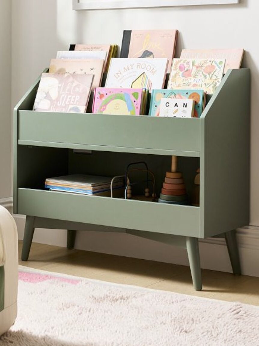 A green kids' bookshelf with picture books next to a cream chair with green cushions. There's a wall shelf with a cat-shaped pillow and framed art on the wall.