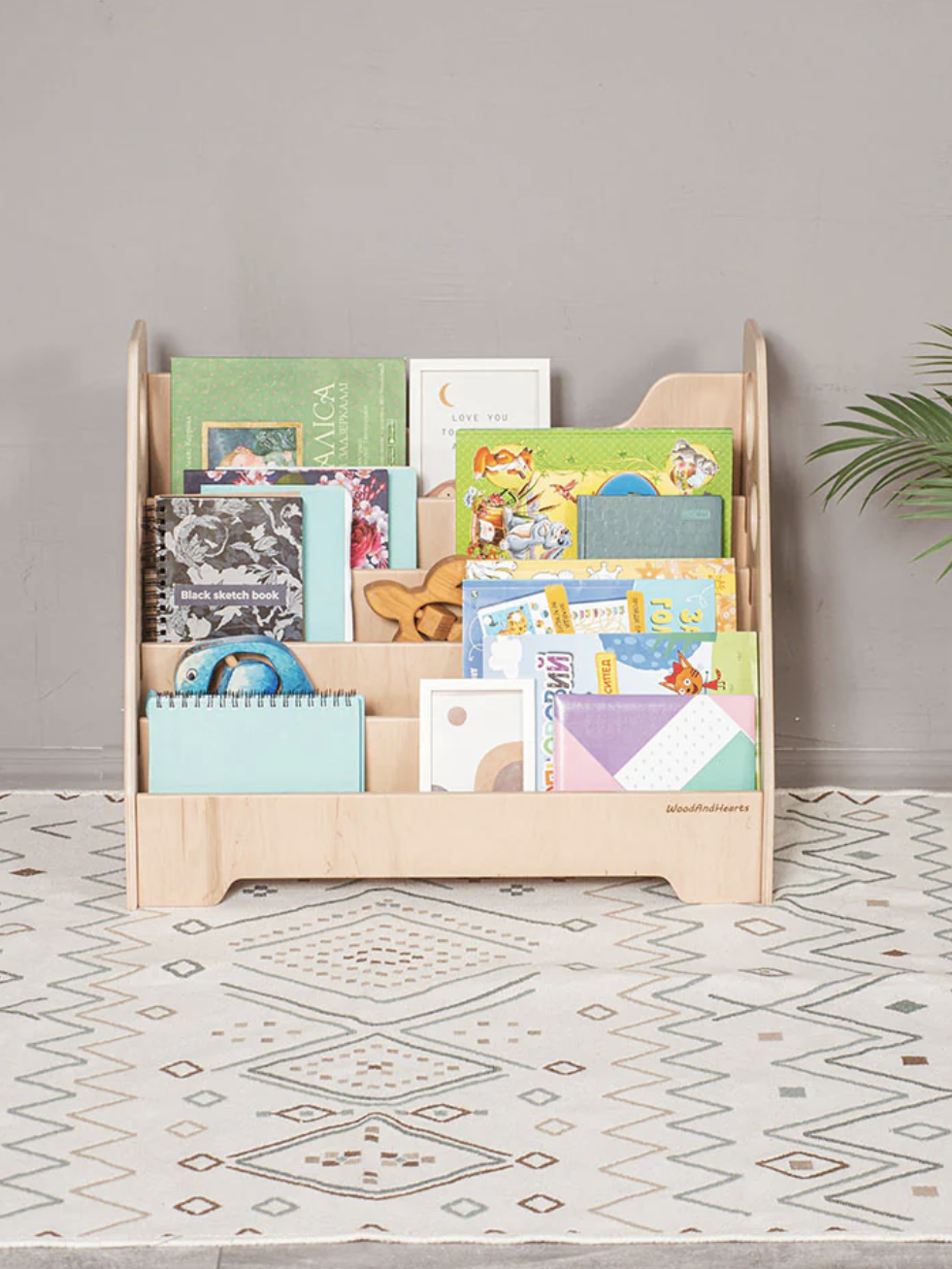 A wooden bookshelf filled with children's books and a toy. It stands on a patterned rug with a plant nearby.