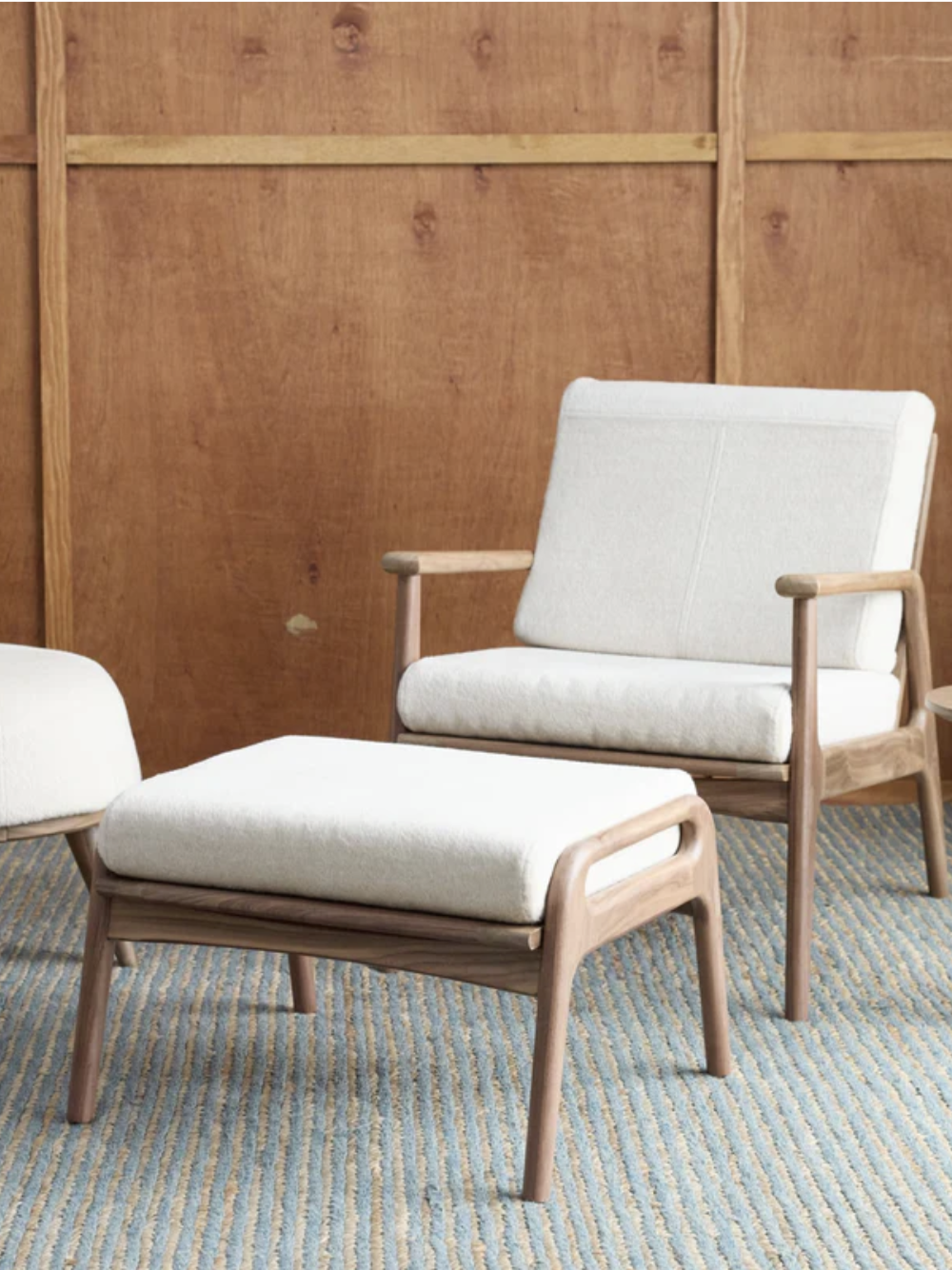 A wooden armchair with white cushions, a matching footstool, and an ottoman are placed on a textured rug in front of a wooden wall. A leafy plant is on the left side.