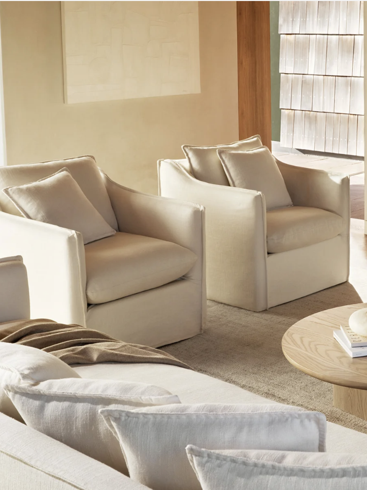 A bright living room with two beige armchairs, a matching sofa, a round wooden coffee table, and minimalist decor. Light flows in through sheer curtains.