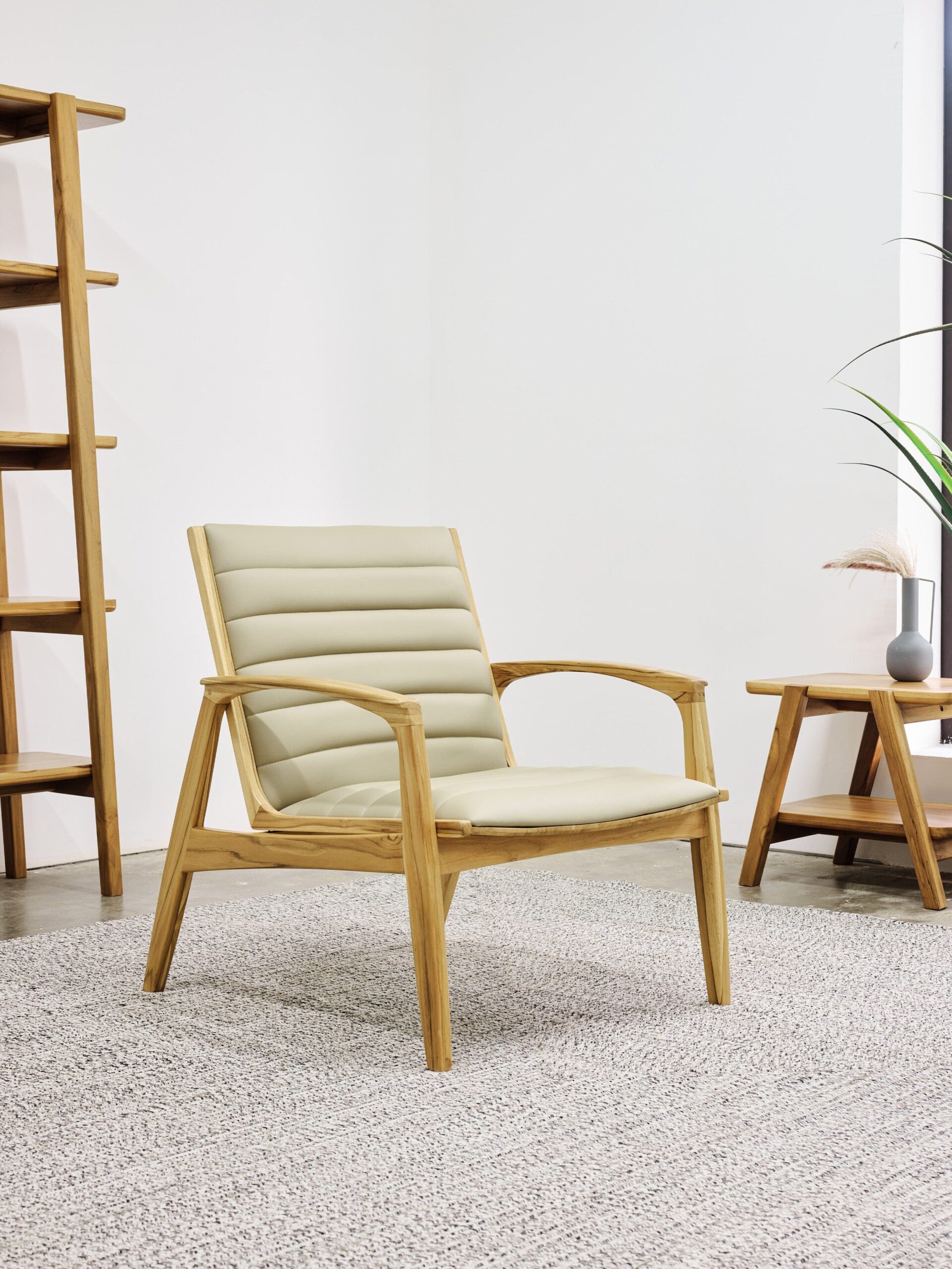 A wooden chair with beige cushioned back and seat is placed on a gray rug in a minimalist room with a wooden shelf and small table.