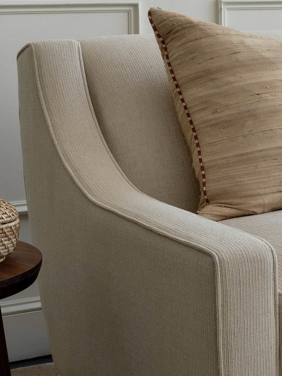 A beige upholstered sofa with a large tan pillow sits next to a wooden side table, which has a woven basket and a vase on top.