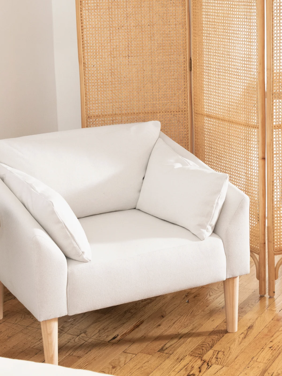 A white cushioned armchair beside a wooden side table with a mug and notebook, near a wicker room divider on a hardwood floor.