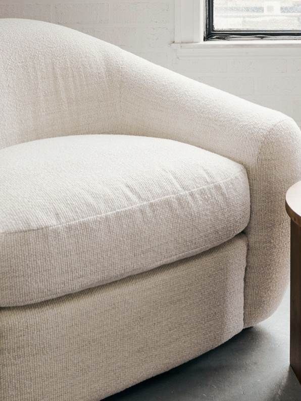 A beige, textured armchair is next to a wooden round table with a glass of tea and a small stack of books on it, set in a room with white brick walls and natural light.