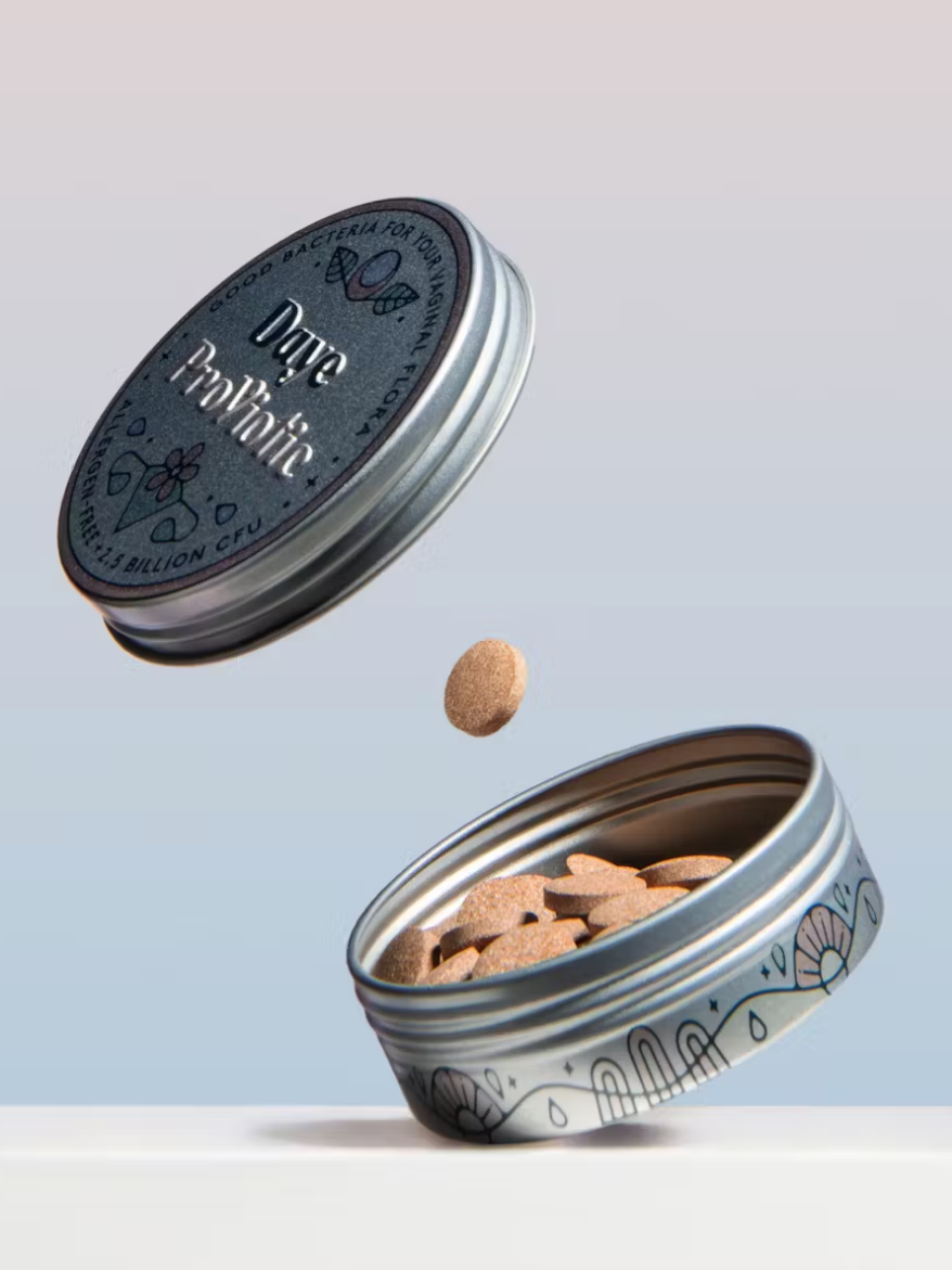 A tin container labeled "Daily Probiotic" is open with multiple tablets inside. One tablet is suspended in the air above the tin.