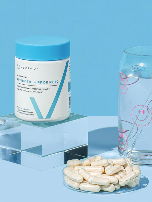 A jar of probiotic supplements on a block beside a glass of water with smiley faces and a straw. Capsules are spilled out on a small white dish in front.