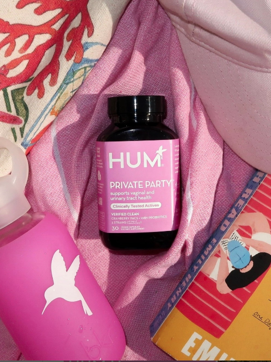 Pink supplements bottle with "HUM" brand lies on pink fabric, surrounded by a pink water bottle, a pink cap, a book, and a colorful patterned item.