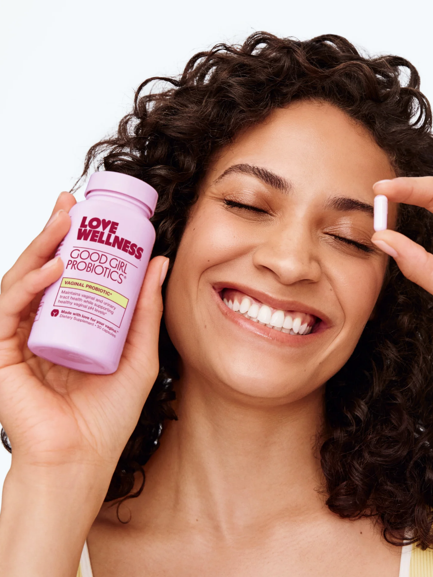 A person smiles while holding a bottle of Love Wellness Good Girl Probiotics and a capsule in hand.