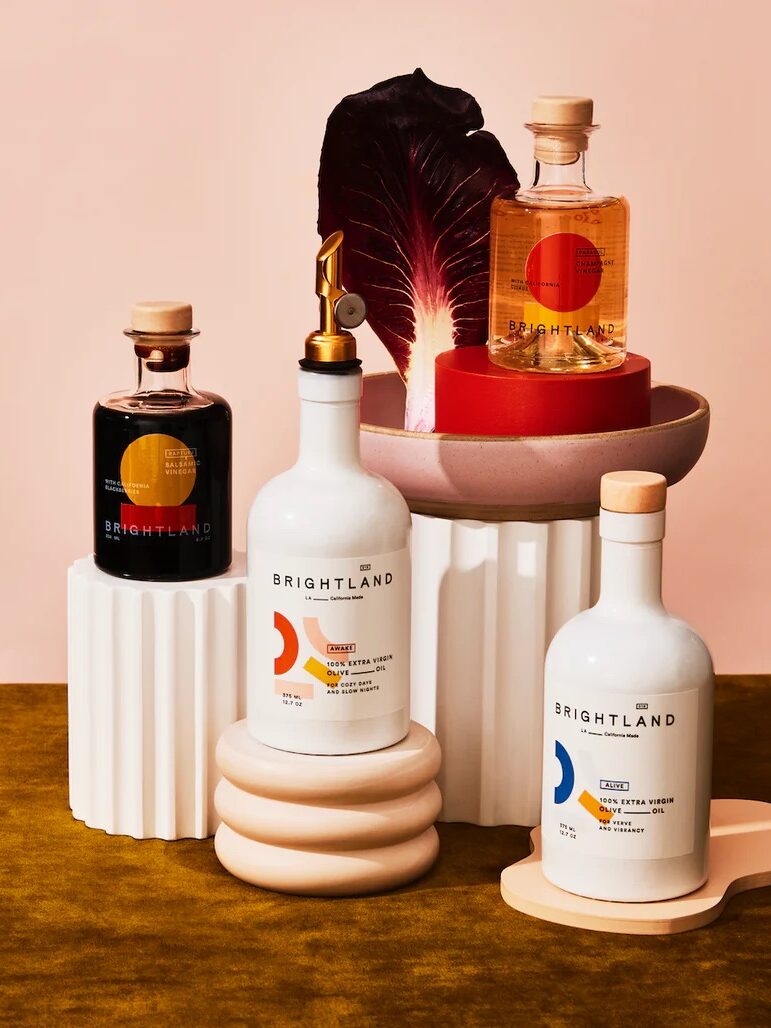 A variety of Brightland bottles, including oils and vinegars, displayed on stacked white and pink platforms with a radicchio leaf in the background.