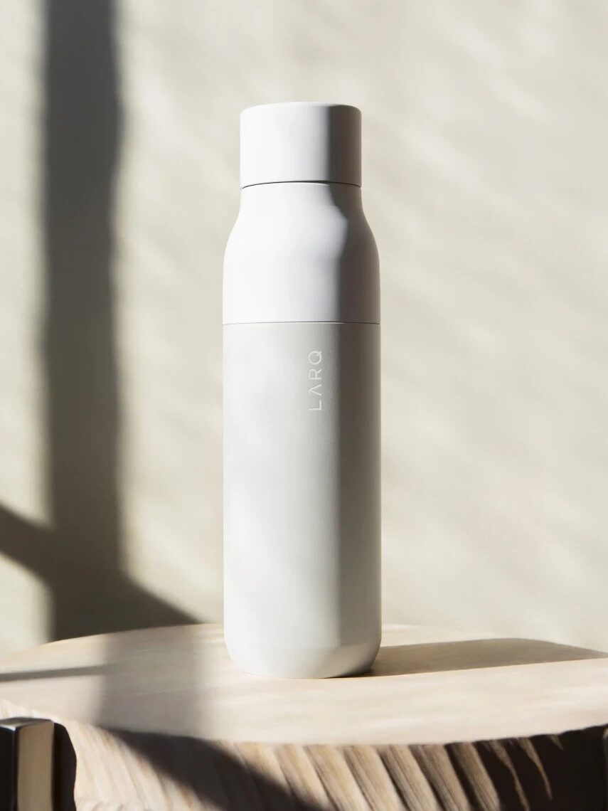 A white water bottle labeled "LARQ" stands on a wooden surface, with sunlight casting shadows in the background.