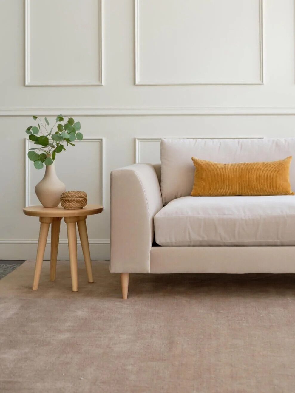 A beige sofa with a mustard yellow pillow is next to two light wooden side tables with a vase and greenery, set against a white paneled wall.