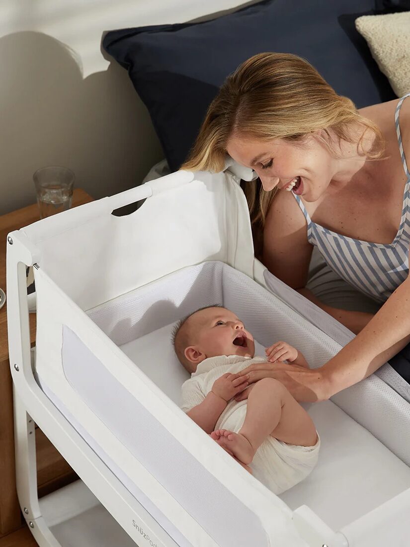 A woman smiles at a baby lying in a white crib next to a bed with a lamp and glass on the nightstand.