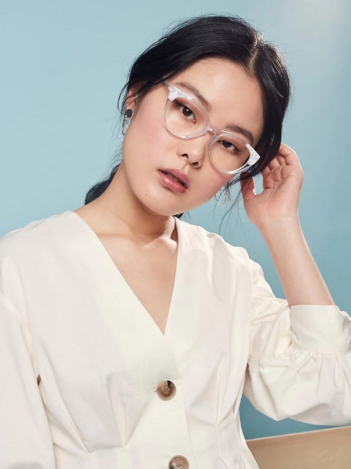 A person with glasses and long hair wears a cream blouse against a light blue background, tilting their head slightly and touching their ear.