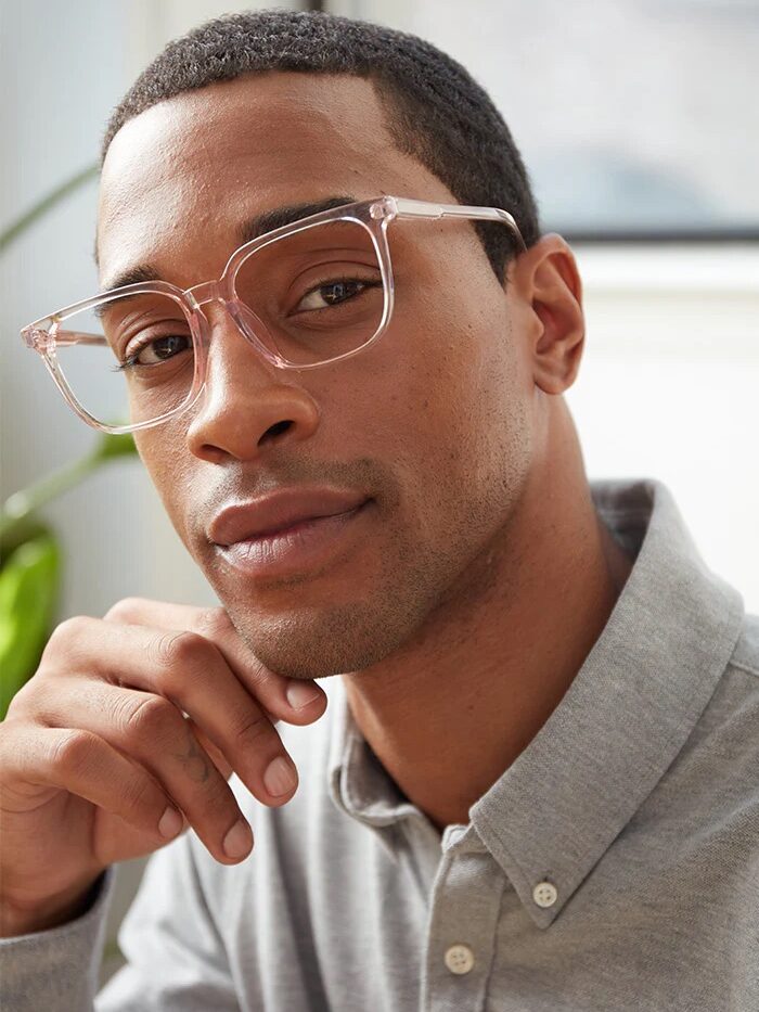 A person wearing transparent glasses and a gray button-up shirt gazes into the camera with a relaxed expression, resting their chin on their hand. A plant is in the background.