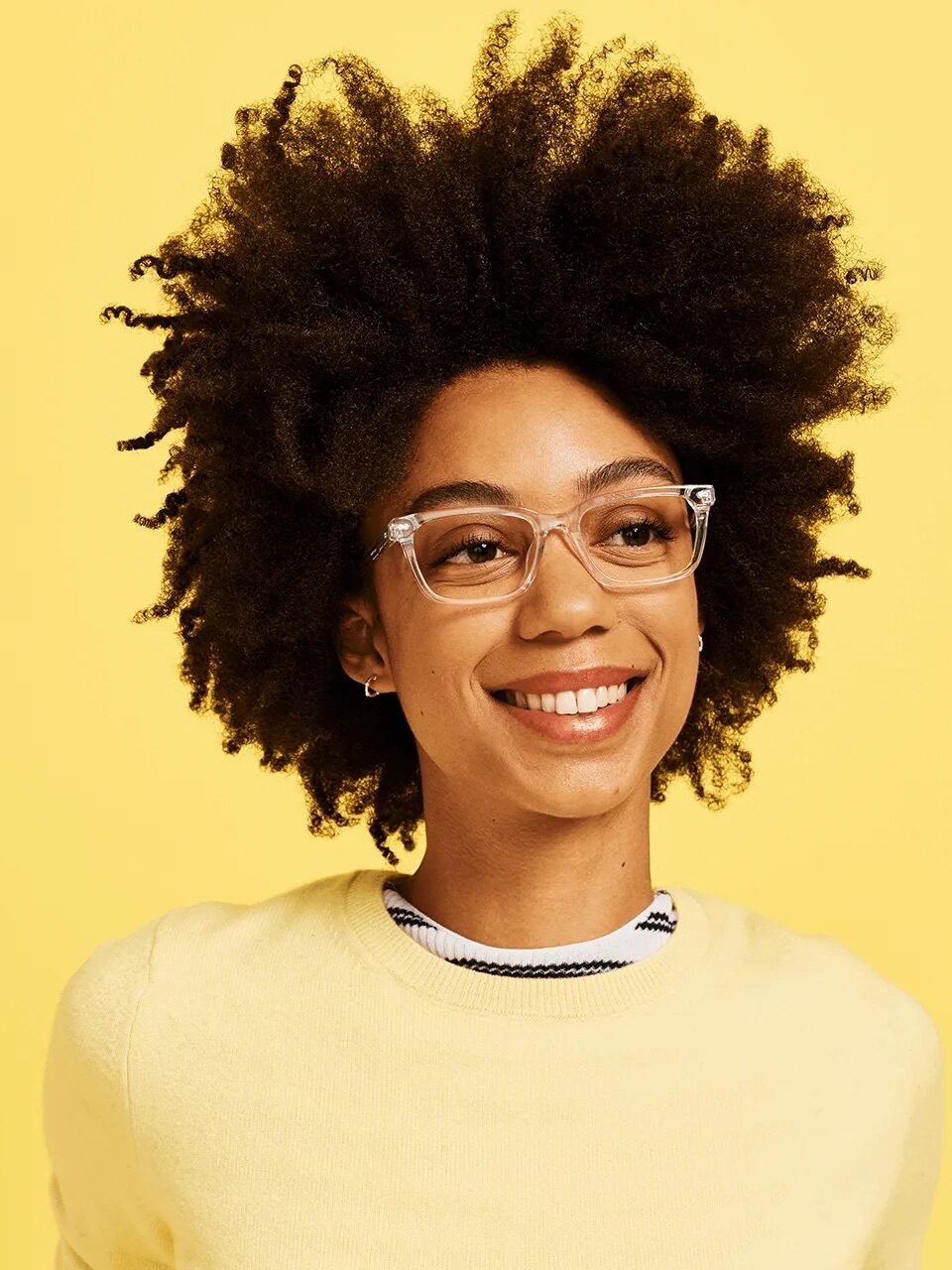 Person with glasses and curly hair smiles against a yellow background.