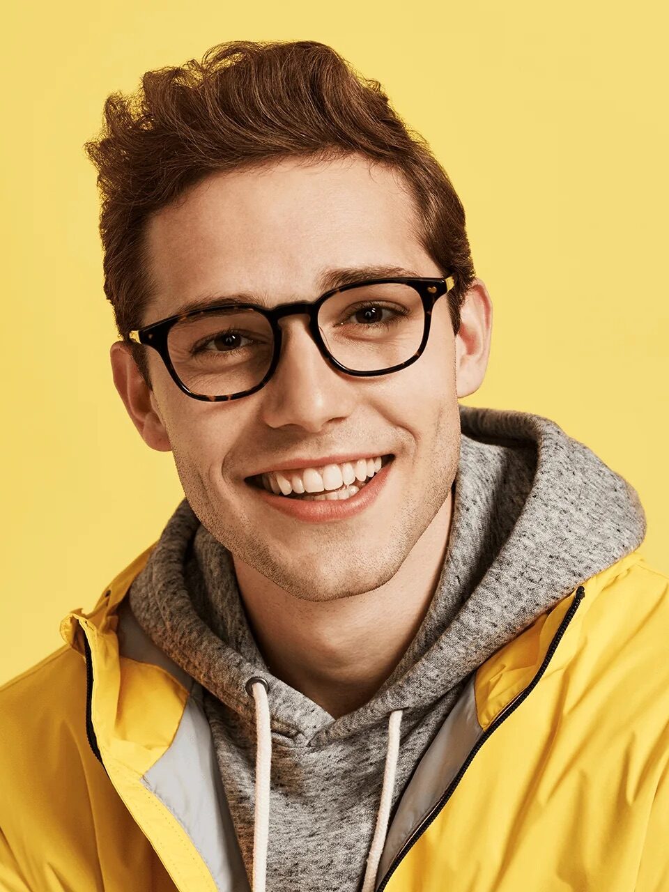 A person wearing glasses, a yellow jacket, and a gray hoodie smiles against a yellow background.