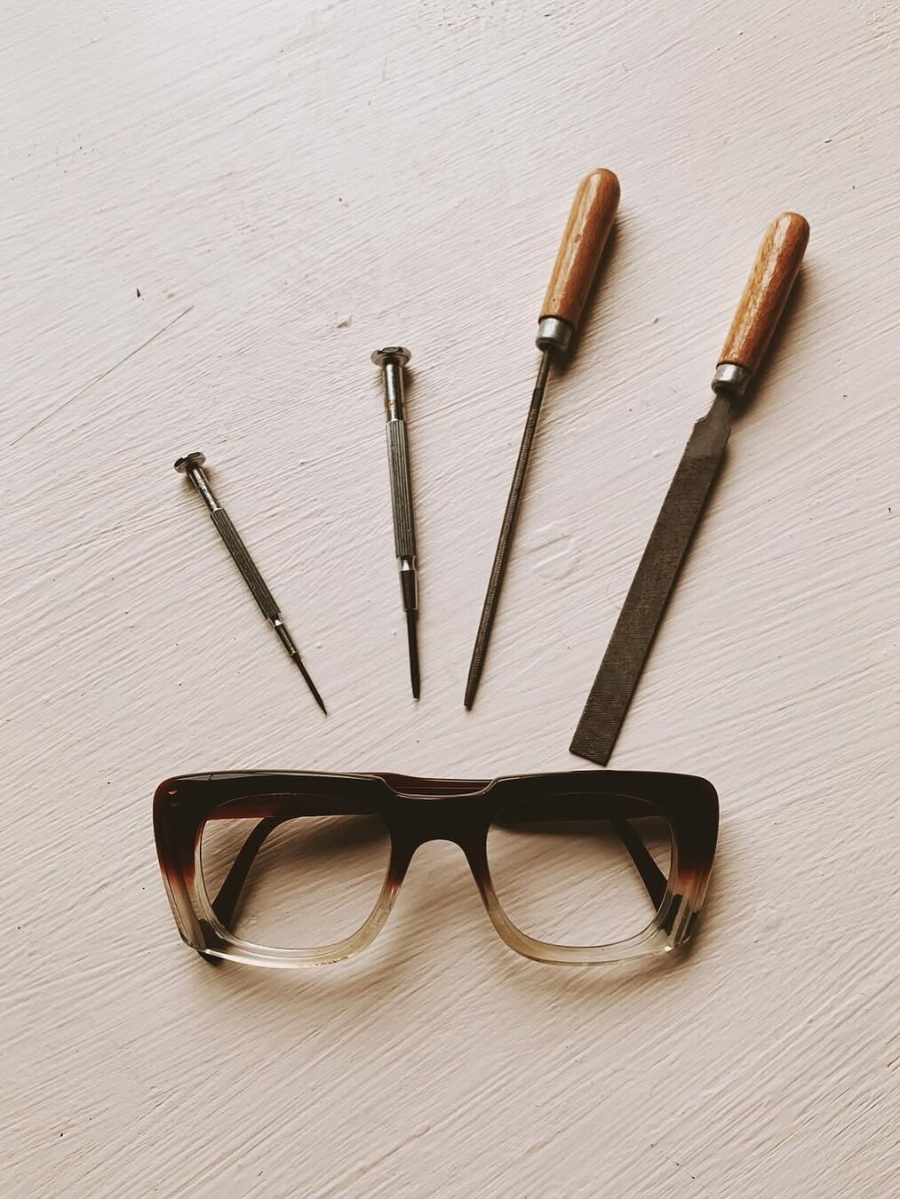 A pair of glasses with two small screwdrivers, a file, and a third tool arranged above them on a white surface.