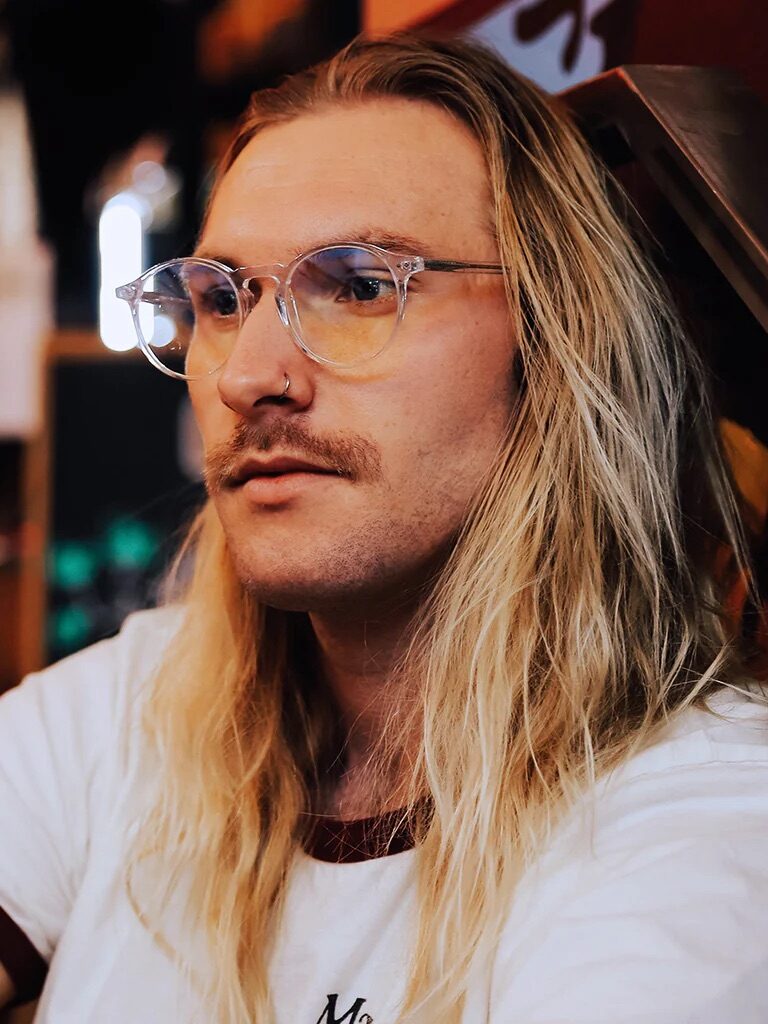 A person with long blonde hair and glasses sits indoors, wearing a white shirt with red trim.