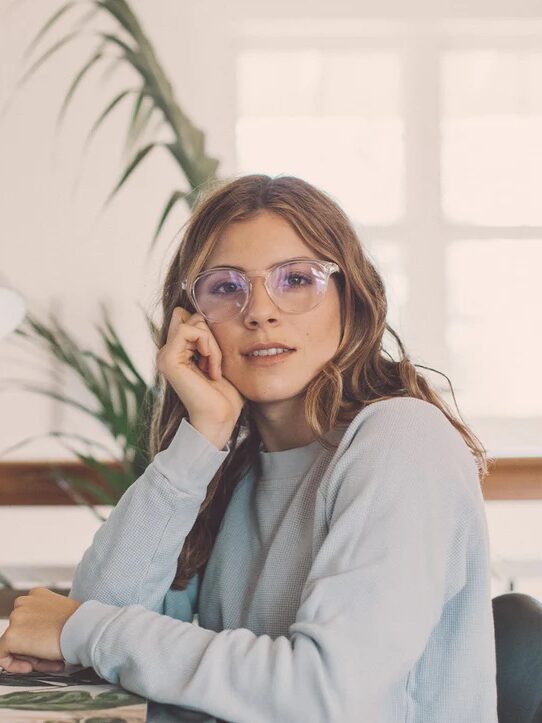 A woman in glasses and a light sweater sits at a desk, hand on chin, looking at the camera. A lamp and plant are in the background.