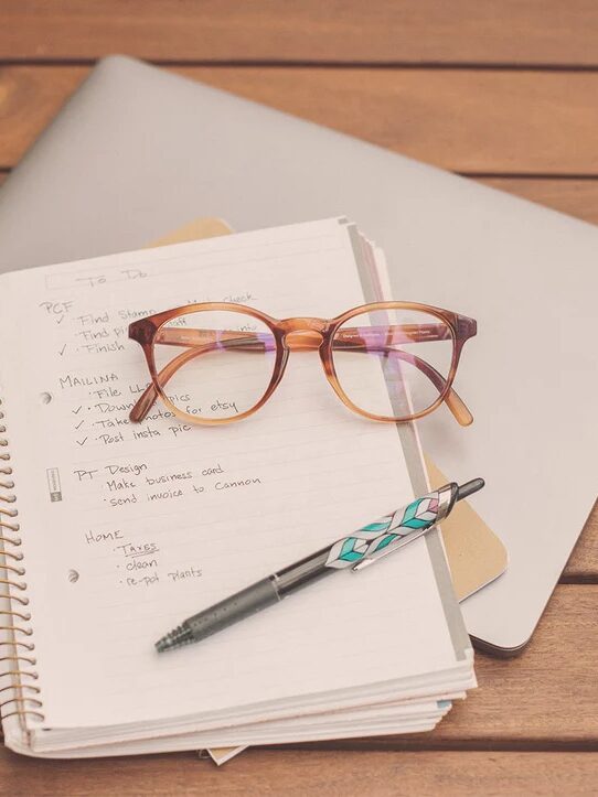 Glasses and a pen on an open notebook with handwritten notes, placed on a closed laptop on a wooden table.