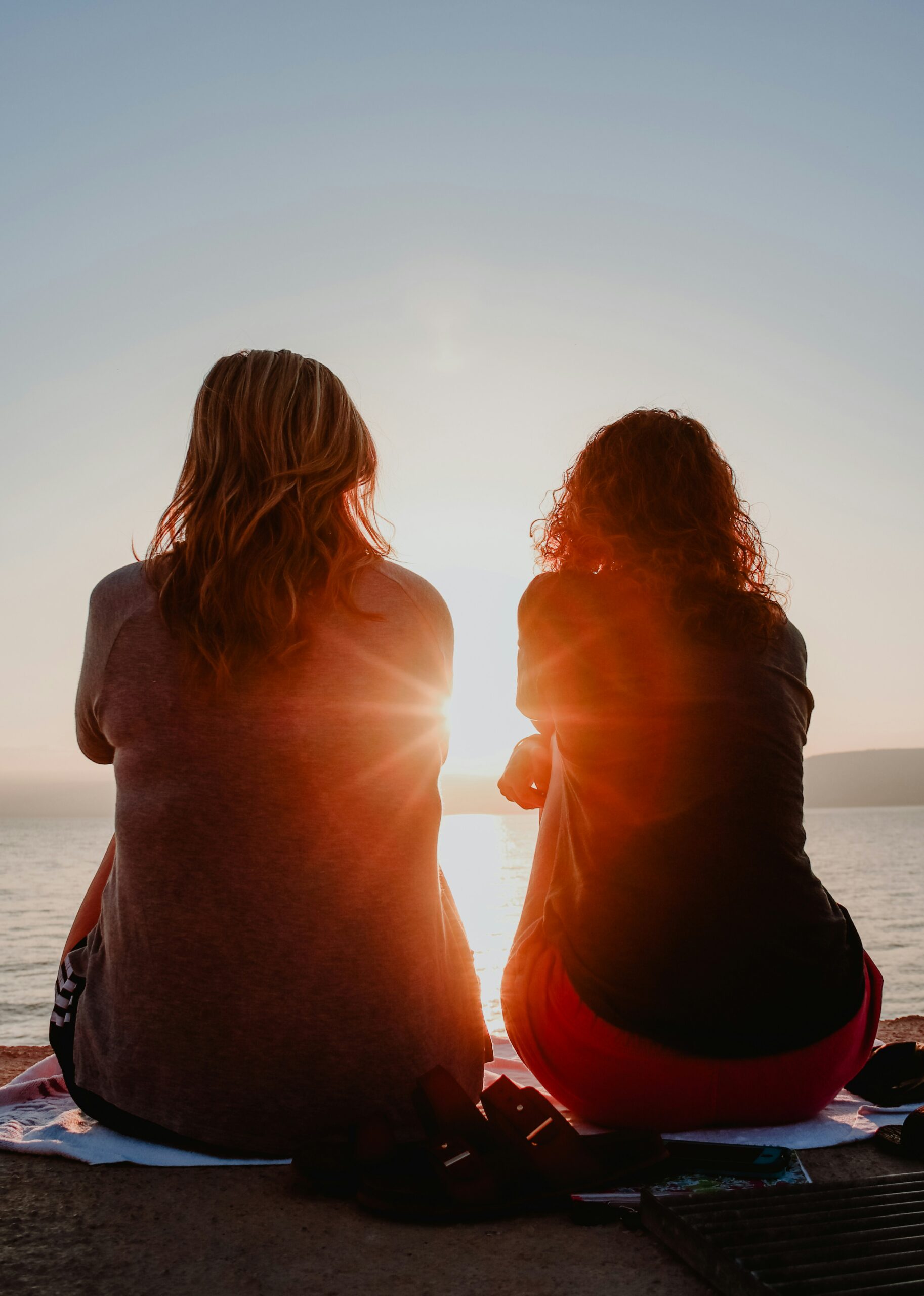 Two people sit on a waterfront, facing the sunset, with sunlight flaring between them.