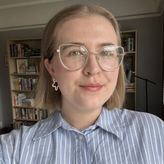 Person with short hair and glasses wearing a striped shirt, standing indoors with bookshelves in the background.