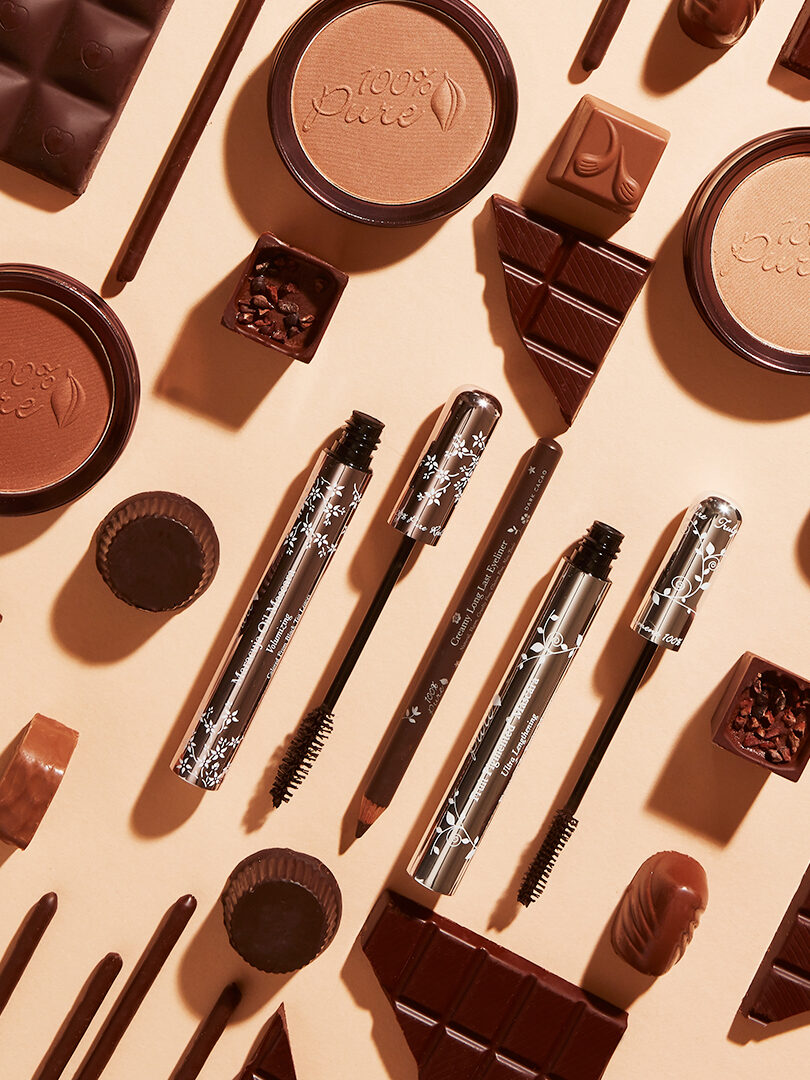 An arrangement of makeup products and chocolate pieces on a beige background, featuring mascaras, brow pencils, and bronzers among various chocolate bars and treats.