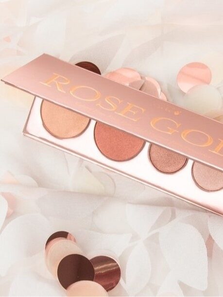 A rose gold eyeshadow palette with five shades is open on a soft white fabric, surrounded by circular confetti.